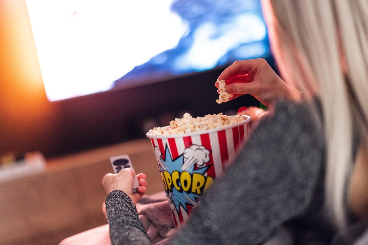 Person reaching into a red-and-white striped popcorn bucket while holding a remote in a dim movie theater.
