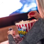 Person reaching into a red-and-white striped popcorn bucket while holding a remote in a dim movie theater.