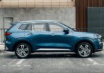 Side view of a blue SUV parked in a paved parking lot beside a curb and modern building façade behind it, showing chrome trim and alloy wheels.