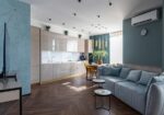 Open-plan living space with a quilted light blue sofa, round nesting tables, and a modern kitchen along the back wall.
