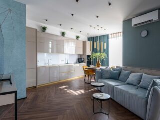 Open-plan living space with a quilted light blue sofa, round nesting tables, and a modern kitchen along the back wall.