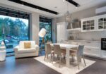 Open-plan modern kitchen and living area with white furnishings and large windows showing trees outside.