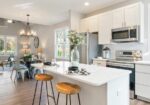 Open-concept kitchen with white cabinets, stainless appliances, and a marble island with wooden stools and a vase of greenery in the foreground.