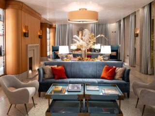 Luxurious hotel lounge with a blue sofa, red and beige cushions, cream armchairs, and a glass coffee table centered by a tall floral arrangement in warm lighting.