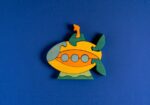 Colorful wooden puzzle-piece toy shaped like a submarine with orange and yellow bodies and three circular portholes on a blue background.