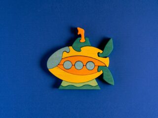 Colorful wooden puzzle piece shaped like a yellow-orange submarine with three portholes on a blue background.